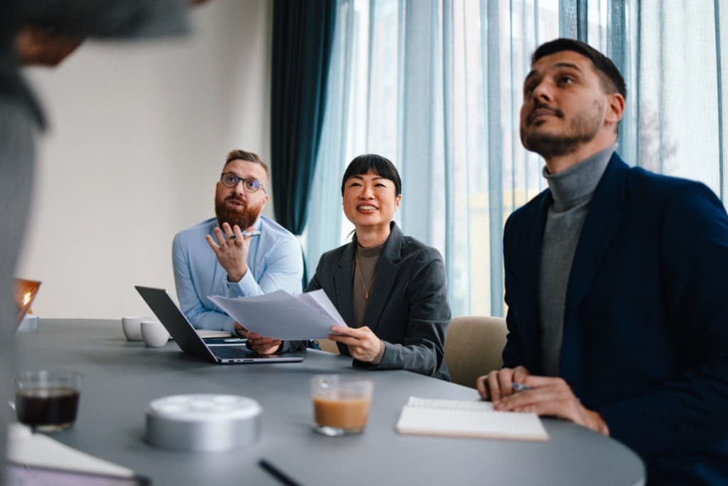 Professionals in discussion around a table in a light-filled wood-paneled office, seen through glass—quiet, focused collaboration.